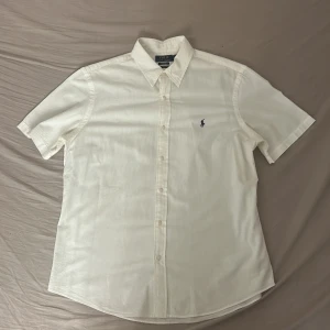 Vit kortärmad skjorta Polo Ralph Lauren - Snygg vit skjorta från Polo Ralph Lauren i bomull med diskreta ränder och klassisk krage. Kortärmad modell med blå broderad logga på bröstet och knäppning framtill. Custom fit för en modern passform.