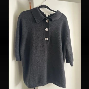 Svart stickad polotröja - Svart stickad polotröja med tre dekorativa pärlknappar framtill och klassisk krage. Tröjan har trekvartsärm och en lite loose passform, perfekt för lager på lager. Enkel men ändå elegant stil som passar till många outfits.