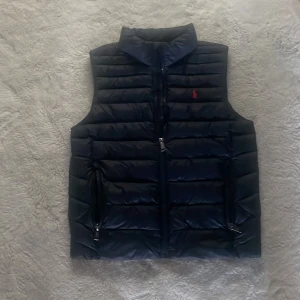 Blå dunväst från Polo Ralph Lauren - Snygg blå dunväst från Polo Ralph Lauren med quiltad design och hög krage. Västen har två fickor med dragkedja och en tydlig Polo-logga broderad i rött på bröstet. Perfekt för lager-på-lager och enkel att matcha till olika outfits.