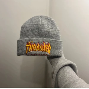 Grå Thrasher mössa, inte använd - Säljer en grå mössa från Thrasher med ikonisk broderad logga i gult och rött med flames på uppvikt kant. Klassisk, enkel passform och stickad i mjukt material. Perfekt för dig som gillar streetwear och skatekultur.