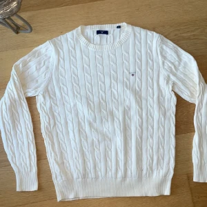Vit kabelstickad tröja från GANT - Vit kabelstickad tröja från GANT med rund halsringning och diskret broderad logga på bröstet. Tröjan har långa ärmar och ribbade muddar vid ärmslut och nederkant. Perfekt för dig som gillar klassisk och stilren look.