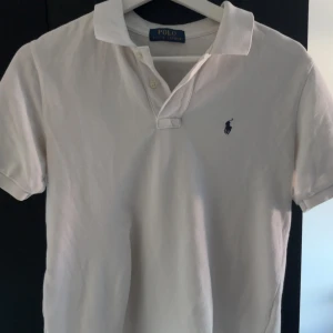 Vit pikétröja från Polo Ralph Lauren - Klassisk vit pikétröja från Polo Ralph Lauren med korta ärmar och ikonisk broderad logga på bröstet. Tröjan har krage och två knappar vid halsen. Skön passform i mjuk bomull, perfekt till jeans eller shorts.