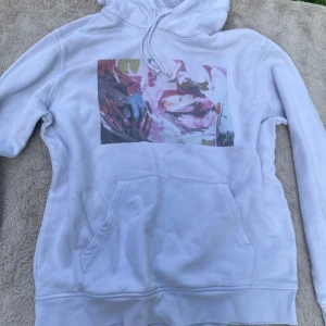 Vit hoodie med konstnärligt tryck - Vit hoodie i storlek L med ett stort, färgstarkt konstnärligt tryck på bröstet. Tröjan har huva med snörning och en klassisk känguruficka framtill. Perfekt för dig som gillar unika och kreativa plagg med streetkänsla.