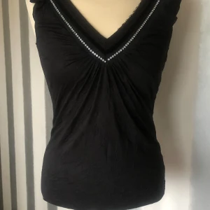 Svart topp med strass från ONLY - Svart ärmlös topp från ONLY med v-ringning dekorerad med strass och spetsdetaljer. Toppens tyg har en lätt skrynklig struktur och smickrande rynk vid bysten. Perfekt för dig som gillar lite extra bling och snygga detaljer.