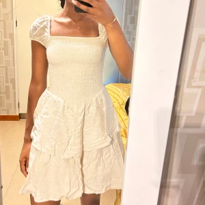 kortklänning med puffärm - Supersöt ljus kortklänning med smockad överdel, puffärmar och volangig kjol. Fyrkantig ringning både fram och bak ger en trendig vibe. Endast testad