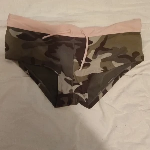 Bikini nederdel / shorts  - Snygga badshorts med camouflage-mönster i gröna och bruna toner, och en ljusrosa midja med snörning. Perfekta för stranden eller poolen när du vill sticka ut lite extra. Tight passform och sportig look.