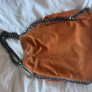 Orange axelväska från Stella McCartney - Cool orange axelväska från Stella McCartney med grova silverfärgade kedjor längs kanterna och som axelrem. Väskan är i mjuk mocka och har magnetlås. Insidan är fodrad med logotypmönstrat tyg och har en innerficka med dragkedja. 