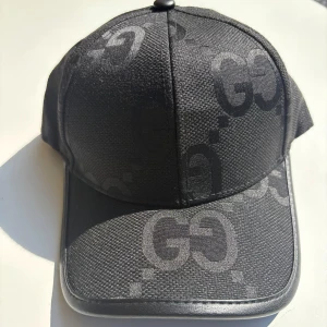 Svart keps från Gucci med GG-mönster - Snygg svart keps från Gucci med ikoniskt GG-mönster ton-i-ton över hela kepsen. Kepsen har böjd skärm med läderdetalj längs kanten och justerbar rem bak i läder. Tillverkad i slitstark polyester med exklusiv känsla.