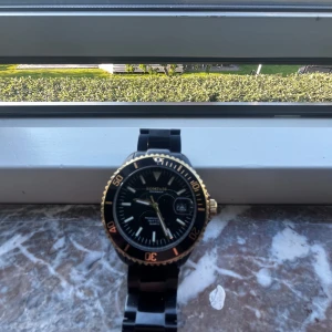 Svart och guldig klocka från Kompass ny pris 3400 - Stilren svart klocka från Kompass med guldig vridring och detaljer. Klockan har svart länkarmband och tydlig urtavla med datumvisning. Vattentät upp till 5 ATM och passar perfekt för dig som gillar en klassisk men ändå modern look.