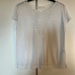 Vit topp med spetsdetalj från ONLY - Vit topp från ONLY i storlek XS med korta ärmar och rund hals. Baksidan har en snygg spetsdetalj i triangelform som ger en cool och feminin touch. Perfekt till jeans eller kjol för en fräsch look.