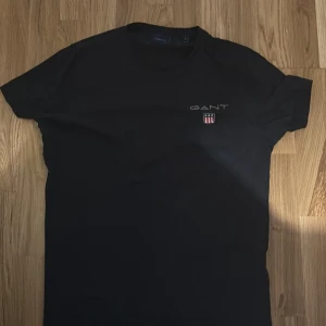 Svart GANT t-shirt med logga - Svart t-shirt från GANT med klassisk rund hals och korta ärmar. På bröstet finns GANT-loggan och ett broderat märke i rött, vitt och blått. T-shirten är i mjuk bomull och har en clean, stilren look som funkar till allt.