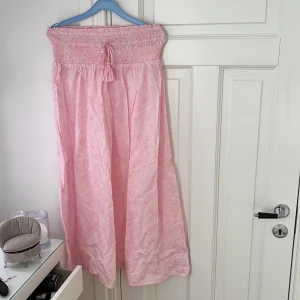 Ljusrosa långkjol från Gina Tricot - Supersöt ljusrosa långkjol från Gina Tricot med smock i midjan och knytband med tofsar. Kjolen har ett diskret paisleymönster och är gjord i ett tunt, luftigt bomullstyg. Perfekt för varma dagar när du vill känna dig både chill och stylish.