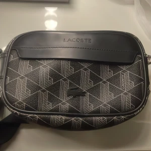 Svart axelväska från Lacoste - Snygg svart axelväska från Lacoste med geometriskt vitt mönster och präglad logga framtill. Väskan har justerbar rem och dragkedja upptill. Tillverkad i slitstarkt syntetmaterial med stilren design och diskreta detaljer.