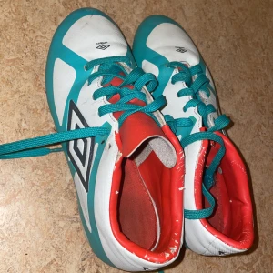 Turkosa och vita Umbro fotbollsskor - Säljer ett par snygga fotbollsskor från Umbro i turkos, vitt och rött. Skorna har snörning, röda dobbar och en cool design med Umbro-logga på sidan. Insidan är röd och ovandelen är i syntetmaterial. Perfekta för dig som vill sticka ut på planen! Köpte de innan för 600kr det vara 5månader sen