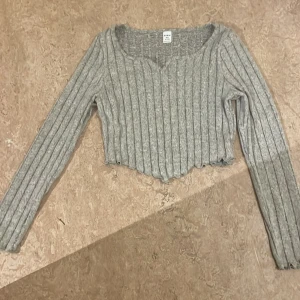 Grå croppad ribbad stickad tröja - Grå croppad stickad tröja från SHEIN med breda ribbade ränder och v-ringad hals. Långärmad modell med vågig kant nertill som ger en trendig look. Perfekt att styla med höga jeans eller kjol. 12/13 år men är liten i storleken! Aldrig använd!