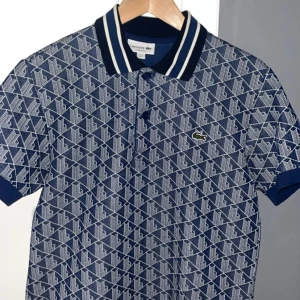 Blå mönstrad Lacoste piké - Snygg blå pikétröja från Lacoste med vitt geometriskt mönster och klassisk krage med vita ränder. Ikoniska krokodilloggan på bröstet och knäppning framtill. Perfekt för dig som gillar sportig och stilren vibe.