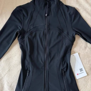 Svart träningsjacka från Lululemon - Snygg svart träningsjacka från Lululemon med figurnära passform och hel dragkedja framtill. Jackan har långa ärmar, två dragkedjefickor och diskret logga på bröstet. Perfekt för dig som vill ha en sportig och stilren look.