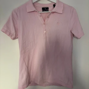 Ljusrosa pikétopp från Gant - Säljer en ljusrosa pikétopp från Gant i premium bomull. Klassisk krage, knäppning med tre knappar och korta ärmar. Diskret broderad logga på bröstet. Perfekt för en preppy och fräsch look. Nypris 1000kr