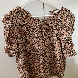 Blommig blus i svart och rosa - Supersöt blommig blus med svart botten och små blommor i rosa, gult och vitt. Blusen har puffiga trekvartsärmar och rund halsringning. Perfekt för dig som gillar romantiska och färgglada plagg.