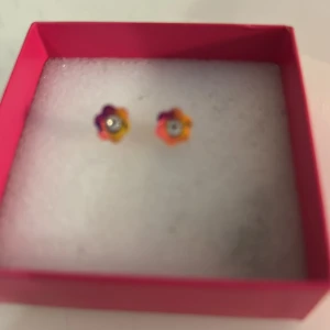 Färgglada blommiga örhängen - Söta små örhängen i form av blommor med flera färger som orange, gul, rosa och lila. De har en liten glittrande sten i mitten som ger extra bling. Perfekta för dig som vill ha något lekfullt och färgstarkt i öronen.