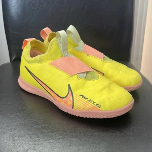 Nike Mercurial Air Zoom fotbollsskor - Nike Mercurial Air Zoom fotbollsskor i neon-gult med rosa detaljer och sula. Skorna har stor AIR-logga på sidan, elastiskt band över vristen och dragflikar i rosa och vitt. Ovandelen är i syntetmaterial och sulan är mönstrad för bra grepp på inomhusplan.