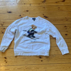 Vit  Polo Ralph Lauren troja - Vit sweatshirt från Polo Ralph Lauren med ikoniska Polo Bear som åker skidor tryckt på bröstet. Tröjan har rund hals, långa ärmar och ribbade muddar. Perfekt för dig som gillar klassisk stil med en twist. Materialet är mjukt och skönt bomull.