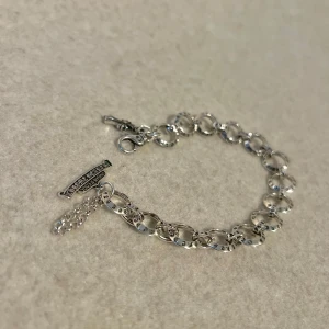 Rockstar Charm Bracelet with Crosses - Ett kraftfullt armband med en kombination av olika berlocker, inklusive kors och inskriptioner på varje länk. Armbandet är 19,5 cm långt med en 1,5 cm förlängning, vilket gör att du kan justera det för en perfekt passform. Den unika designen ger ett edgy och distinkt uttryck, perfekt för att addera en rockig touch till din stil. 