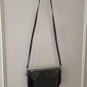 Svart axelväska från Carin Wester - Snygg svart axelväska från Carin Wester i glansigt skinnliknande material. Väskan har justerbar axelrem, silverfärgade metalldetaljer och dragkedja upptill. Perfekt storlek för det viktigaste och stilren design som lyfter varje outfit.