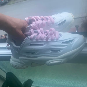 Adidas sneakers med rosa snören - Chunky vita Adidas sneakers med futuristisk design och rosa snören. Skorna har en tjock sula och detaljerade utskärningar på sidorna. Perfekta för dig som vill sticka ut med en cool och modern look.