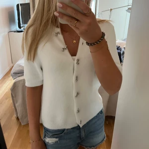 Vit kofta från Zara - Superfin vit kofta från Zara med korta ärmar och söta rosettformade knappar framtill. Väldigt mjukt och skönt material. Ribbad kant nedtill och vid ärmslut. Väligt liten i storlek.