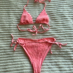 Rosa bikini med smock och knyt - Supersöt rosa bikini med smockat tyg och knyt i både topp och trosa. Får en att se jätta tan ut till o me om man ej är det! Triangelformad överdel med tunna band som knyts i nacken och ryggen. Underdelen har höga ben och knyt i sidorna. Perfekt för stranden eller poolen!