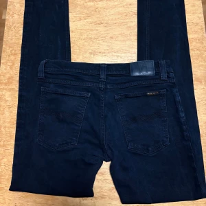 Mörkblå skinny jeans från Nudie - Snygga mörkblå skinny jeans från Nudie Jeans i ekologisk bomull. Klassisk femficksmodell med silverfärgade nitar och diskret broderi på bakfickorna. Jeansen har en smal passform och normal midja, perfekta för en clean och stilren look.