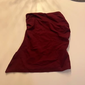Vinröd  tubtopp - Säljer en vinröd bandeau tubtopp med asymmetrisk skärning. Toppen är ärmlös och har en tight passform, perfekt för att skapa en snygg och enkel look. Tillverkad i mjukt bomullsmaterial som känns skönt mot huden.
