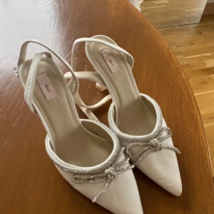 Vita pumps med strassrosett från Nelly - Snygga vita pumps från Nelly med spetsig tå och låg klack. Skorna har en glittrig strassrosett framtill och remmar runt ankeln. Ytan är i mockaliknande material och de är perfekta för dig som vill sticka ut med lite extra bling.