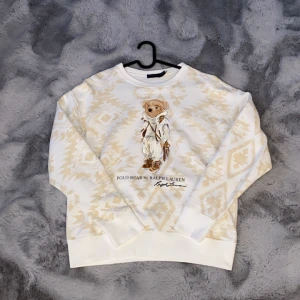 Polo Bear sweatshirt från Ralph Lauren - Vit och beige sweatshirt från Polo Ralph Lauren med ikoniska Polo Bear-trycket framtill. Tröjan har ett snyggt geometriskt mönster i beige och ribbade muddar vid ärmslut och nederkant. Perfekt för dig som gillar streetstyle och klassiska detaljer.