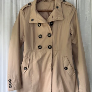 Beige trenchcoat med knappar - Stilren beige trenchcoat med dubbelknäppning och klassiska svarta knappar. Jackan har långa ärmar, krage med axelklaffar och två fickor framtill. Perfekt för dig som vill ha en tidlös och snygg look till vardags.