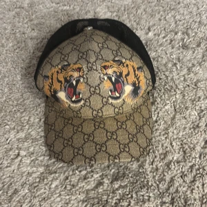 Gucci keps helt oanvänd. - Snygg keps från Gucci med klassiskt GG-mönster i beige och brunt, två stora tigrar framtill och svart mesh baktill. Justerbar rem med grönt och rött band. Perfekt statement-piece för dig som vill sticka ut.
