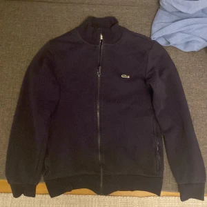 Mörkblå zip från Lacoste XS - Mörkblå zip från Lacoste, storlek XS. Använd endast ett par gånger men säljs pga att den är för liten. Finns i Sthlm