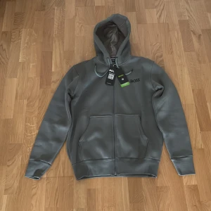 Grå hoodie från Hugo Boss med dragkedja - Snygg grå hoodie från Hugo Boss med dragkedja framtill och två stora fickor. Tröjan har huva med snörning och är tillverkad i mjukt material som känns skönt mot huden. Perfekt för en chill och stilren look.