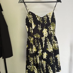 Svart blommig sommarklänning H&M Helt Ny aldrig använd - Supersöt svart kort klänning från H&M med tunna axelband och blommigt mönster i vitt och gult. Smockad rygg för extra komfort och flowy kjol. Perfekt för varma dagar och picknickar. Sitter snyggt och är lätt att matcha med sneakers eller sandaler.