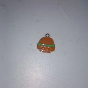 Halsbandshänge i form av hamburgare - Superkul halsbandshänge som ser ut som en hamburgare med brunt bröd, grön sallad och orange fyllning. Perfekt för dig som gillar unika och lekfulla smycken. Passar att hänga på en kedja eller armband för en personlig touch.
