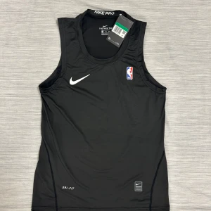 Svart Nike Pro NBA ärmlös träningströja - Svart ärmlös träningströja från Nike Pro med NBA-logga på bröstet och vit Nike Swoosh. Tillverkad i lätt och stretchig polyester med Dri-FIT-teknologi som håller dig torr under intensiva träningspass. Perfekt för basket eller gymmet.
