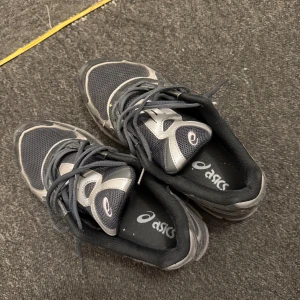 Svarta och grå Asics sneakers - Säljer ett par svarta och grå sneakers från Asics med mesh och syntetdetaljer. Skorna har snörning och en platt sula, perfekt för dig som gillar sportig stil. Klassisk Asics-logga på sidan och bekväm passform.