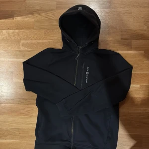 Svart hoodie från Sail Racing - Svart hoodie från Sail Racing med hel dragkedja och bröstficka med dragkedja. Tröjan har huva med snörning och diskret logga på bröstet. Perfekt för dig som gillar sportig och clean stil.