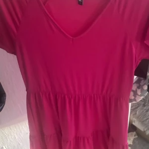 Rosa kortklänning från Pieces - Supersöt rosa kortklänning från Pieces med v-ringning och volangdetaljer. Klänningen har korta ärmar och en luftig passform, perfekt för varma dagar. Tillverkad i mjukt material som känns skönt mot huden.