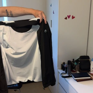 Svart vindjacka från Lacoste och byxor från Lacoste  - Säljer en svart vindjacka från Lacoste med vita ränder längs ärmarna och stor logga på ryggen. Jackan har dragkedja och är tillverkad i lätt polyester, perfekt för dig som gillar sportig och clean stil. Klassisk krokodil på bröstet och elastiska muddar. Samt byxor hela sättet originalet pris ligger på 2.700 har använd byxorna 5 gånger och jackan 2 gånger, (i princip helt nya) PRIS KAN DISKUTERAS VID SNABBAFFÄR