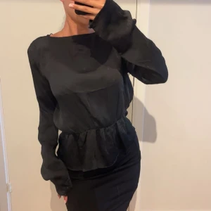 Svart blus med snörning  - Svart blus i glansigt material med lång ärm och snygg snörning i ryggen. Modellen har markerad midja och lätt volang nedtill. Blusen är aldrig använd och köpt på BikBok!
