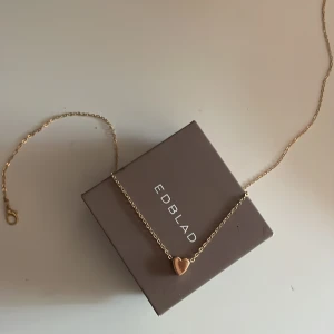 Guldigt halsband med hjärta från Edblad - Snyggt halsband från Edblad i guldton med ett litet hjärta som hänge. Kedjan är tunn och elegant, perfekt för att ge en söt touch till din outfit. Hjärtat är slätt och stilrent, vilket gör halsbandet lätt att matcha med andra smycken. Gammal design och är därför ej i rostfritt stål, pris kan alltid diskuteras 