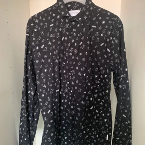 Svart skjorta med julmönster Lindbergh - Svart slim fit skjorta från Lindbergh med vita juliga motiv som snöflingor, granar, paket och renar. Skjortan har långa ärmar och knäppning framtill. Perfekt för dig som vill sticka ut med en rolig och snygg look under vinterns högtider.