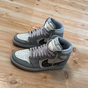 Nike Air Jordan 1 x Dior Highs - Exklusiva Nike Air Jordan 1 x Dior sneakers i grått och vitt skinn med Dior-logga på sidan och genomskinlig sula med Dior-tryck. Hög modell med snörning och perforerad tå för extra stil. Perfekta för dig som vill sticka ut med en ikonisk och lyxig sneaker. Kommer med skosnören och allt som tillgick vid köp så som box osv
